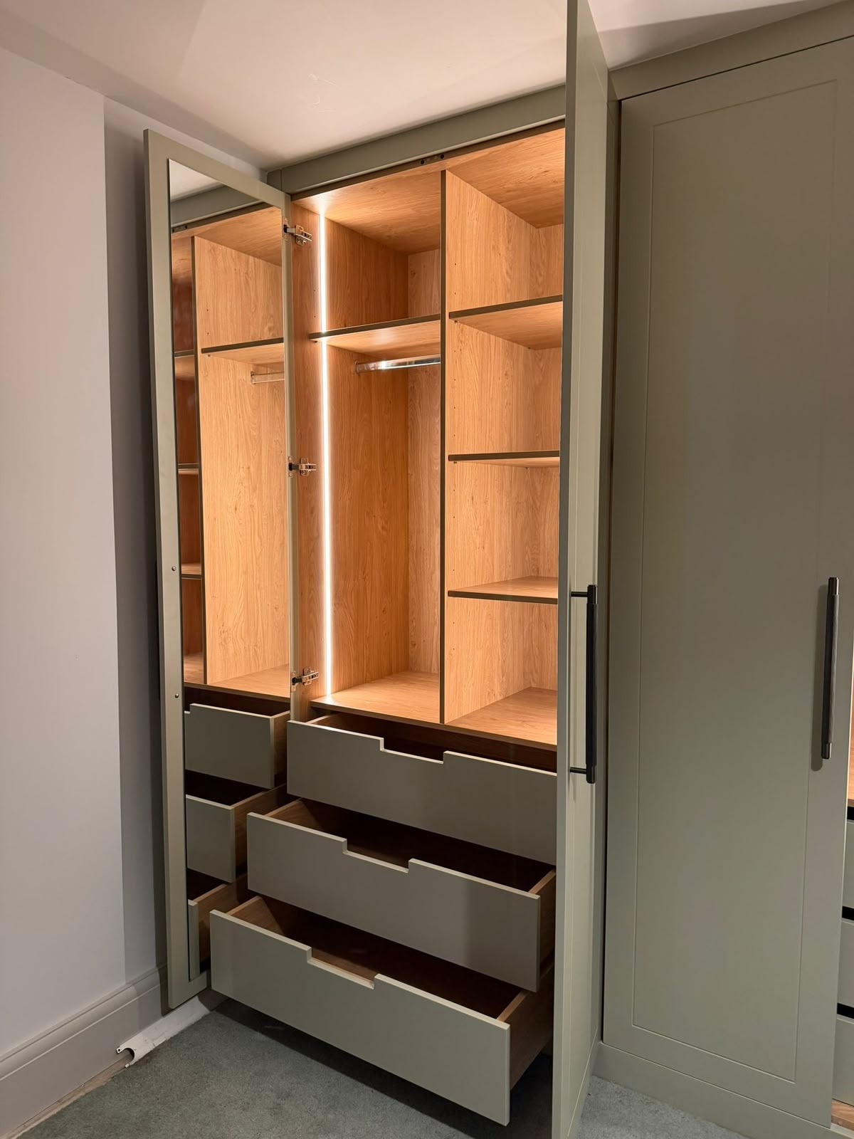 Sage-painted fitted wardrobe with doors open showing oak-lined interior, shelving, hanging rail, deep drawers and integrated LED lighting